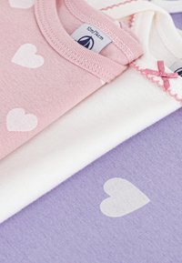 Tri sljedeća pamučna odjeća slojevito: ružičasta s bijelim srcima, bijela i lavanda s srcem. Ima ukrase s valovitim rubovima i vrpcama.