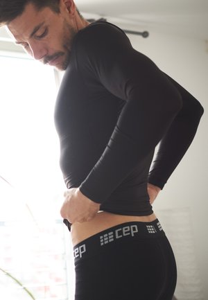 Schwarzes, langärmliges Sportshirt und dunkle Shorts. Glatter, eng anliegender Stoff. 'CEP'-Logo in Grau auf dem Bund, während des Ankleidens getragen.