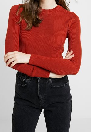 Femme aux longs cheveux bruns portant un pull rouge côtelé ajusté et un jean noir taille haute, debout, les bras croisés.