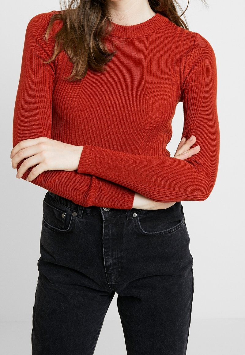 Femme aux longs cheveux bruns portant un pull rouge côtelé ajusté et un jean noir taille haute, debout, les bras croisés.