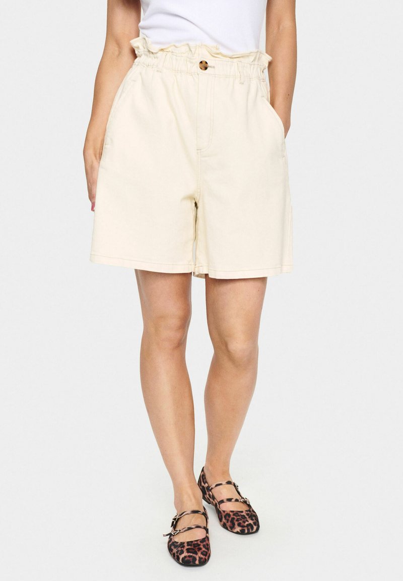 Shorts en coton beige avec une taille froncée, une fermeture à bouton et des poches latérales. Le tissu semble doux avec une texture lisse.