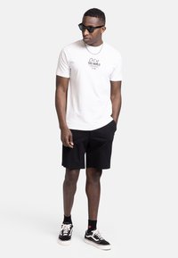 Weißes Baumwoll-T-Shirt mit schwarzem Aufdruck "Out of THIS WORLD", kombiniert mit schwarzen Shorts und schwarz-weißen Turnschuhen. Schwarze Sonnenbrille getragen.