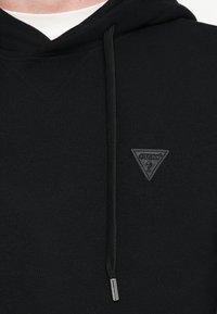 Μαύρο φούτερ με κουκούλα, διαθέτει τριγωνικό δερμάτινο έμβλημα με ανάγλυφο λογότυπο. Το ύφασμα έχει υφή και διαθέτει κορδόνι στη κουκούλα.