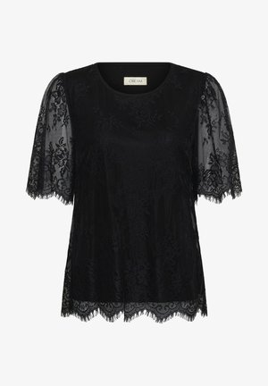 Femme portant un chemisier court en dentelle noire, un jean droit bleu clair et des sandales noires à talons, debout devant un fond clair uni.