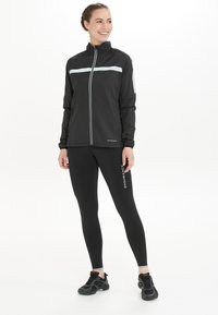Zwarte sportjack met reflecterende strepen, rits aan de voorkant en elastische manchetten, gecombineerd met zwarte leggings met een logo-print en zwarte sneakers.