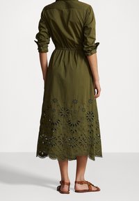 Olivgrön skjortklänning med åtdragen midja, med utskurna blommönster vid fållen. Korta ärmar och krage.