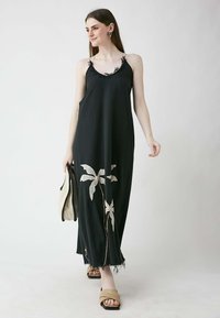 Schwarzes Maxikleid mit Spaghettiträgern, verziert mit Spitze und besticktem weißen Palmenblatt-Design. Strukturierter Baumwollstoff mit fransigem Saum.