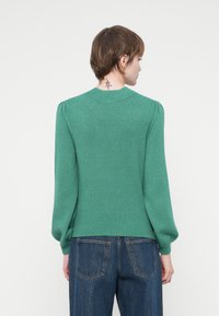 Persona con capelli corti castani e tatuaggio sul collo, che indossa un maglione verde a maniche lunghe e jeans in denim blu, in piedi con la schiena rivolta.