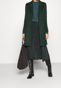 Donkergroene jas, geplooide zwarte rok, blauwe trui, hoge zwarte laarzen met hak, en een zwarte tas. Vlotte texturen en gestructureerde ontwerp elementen.