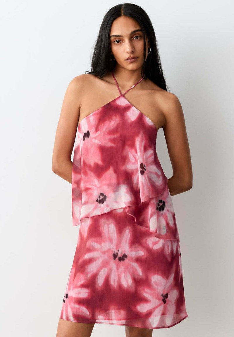 Femme aux longs cheveux foncés portant une robe rose à fleurs sans manches avec un col halter, debout devant un fond gris clair uni.