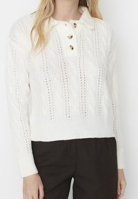 Weißer Pullover mit Zopfmuster, Kragen und drei goldenen Knöpfen sowie gerippten Bündchen. Der gesamte Körper ist mit durchbrochenen Details versehen.