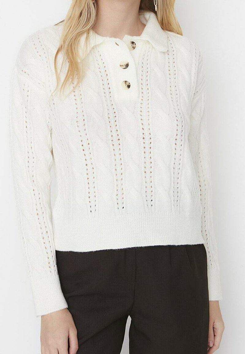 Weißer Pullover mit Zopfmuster, Kragen und drei goldenen Knöpfen sowie gerippten Bündchen. Der gesamte Körper ist mit durchbrochenen Details versehen.