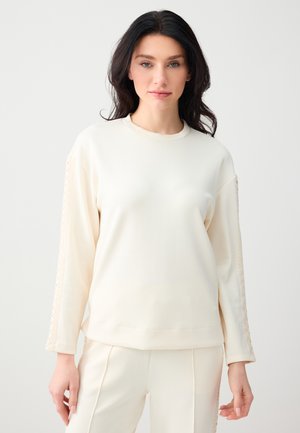 Vrouw met donker haar, gekleed in een crèmekleurige top met lange mouwen en bijpassende broek met kanten details aan de mouwen en zijkanten van de broek, staand tegen een effen achtergrond.