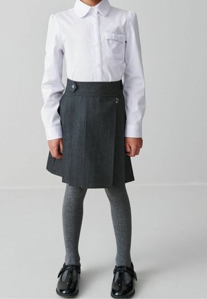 Dziecko ubrane w białą koszulę na guziki, ciemnoszary plisowany spódnicę, szare rajstopy i czarne damskie buty stoi na tle jednolitego tła.