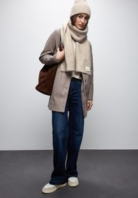 Cappotto beige, berretto e sciarpa in maglia beige, jeans in denim e sneakers beige. Borsa a tracolla marrone con branding minimale. Texture lisce e vestibilità rilassata.