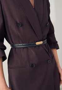Blazer doppiopetto viola scuro con bottoni neri, caratterizzato da una cintura in pelle nera con dettaglio dorato e maniche lunghe.