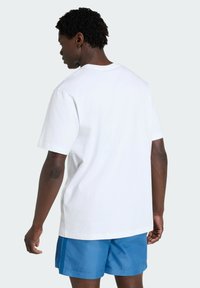T-shirt bianca in cotone a maniche corte, vestibilità comoda, indossata con pantaloncini blu. Il tessuto appare liscio con dettagli di cuciture minimi.