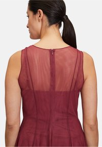 Bordeauxfarbenes Kleid mit einem transparenten Netzrücken, gerundetem Ausschnitt und einem verdeckten Reißverschluss auf der Rückseite. Glattes Material, ärmelloses Design.