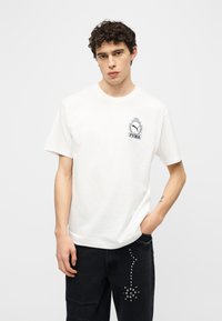 Νέος άνδρας με σγουρά μαλλιά, που φοράει λευκό μπλουζάκι Puma και μαύρο τζιν με καρφιά, στέκεται μπροστά σε ένα απλό φωτεινό φόντο.