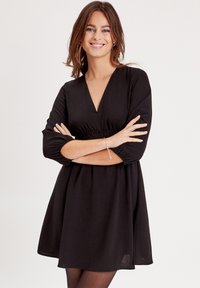 Robe noire avec un décolleté en V, taille froncée et manches bouffantes trois-quarts. Le matériau semble léger avec une texture lisse.