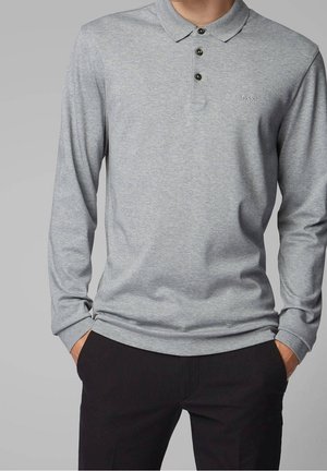 Mann trägt ein hellgraues, langärmeliges Poloshirt mit drei Knöpfen und schwarze Hose, Hände in den Taschen, einfarbiger Hintergrund.