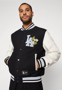 Svart ull varsity-jakke med hvite lær ermer, stripete ribbede mansjetter, og LA-logo broderi med blomsterdetaljer på brystet.