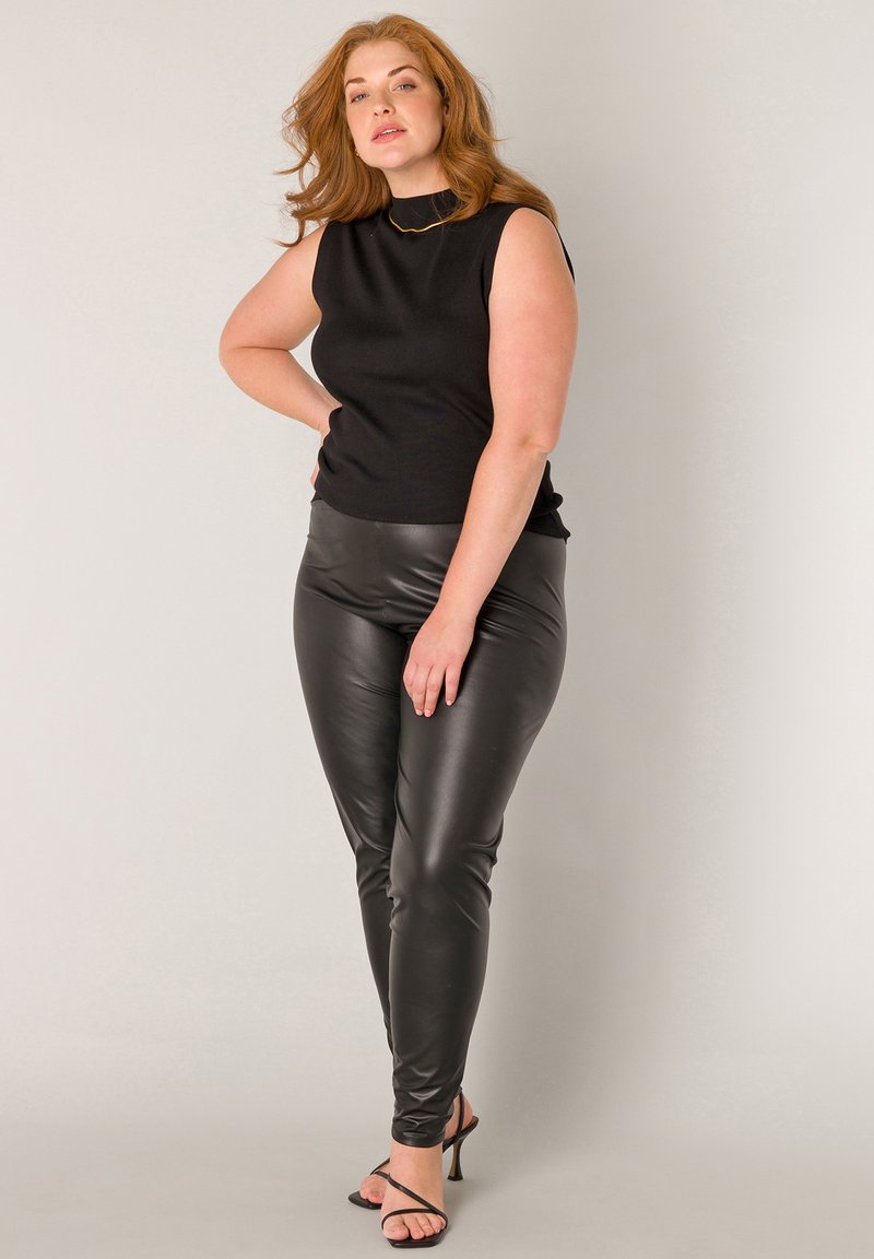 Woman with red hair wearing sleeveless black top, black faux leather pants, and black strappy heeled sandals, standing with hand on hip.