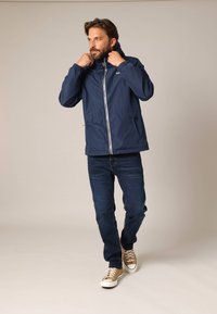 Veste imperméable marine avec capuche, fermeture éclair, deux poches latérales ; portée sur un jean bleu et des baskets beiges, se tenant devant un fond neutre.