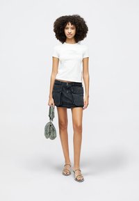 Bílý tričko s krátkým rukávem a bočními rozparky, spárované s černou džínovou mini sukní s předními kapsami. Modelka drží vzorovanou tašku a nosí sandály.