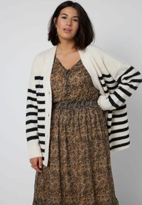 Gestreepte cardigan in crème en navy met chunky breiwerk, knopen aan de voorkant, gecombineerd met een bruine jurk met patroon en een getailleerde taille.