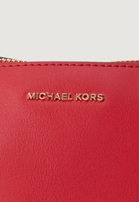 Sarkana ādas dizainera maka ar zelta izspiedtu "MICHAEL KORS" logo, gluda virsma ar redzamām šuvēm un rāvējslēdzēja detaļu.