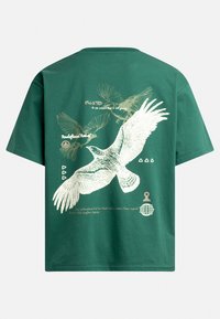 T-shirt vert à manches courtes avec un grand graphisme d'aigle blanc, de petits oiseaux et un texte imprimé en différentes polices et tailles.