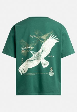 Green cotton t-shirt featuring a large white illustrated bird with outstretched wings, accompanied by printed text and graphic elements in beige.
