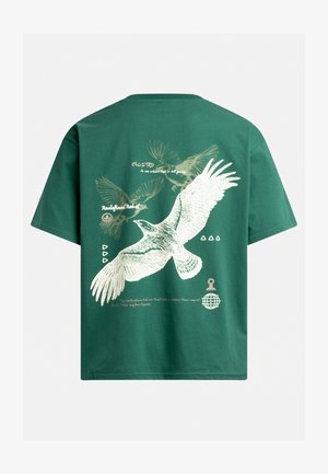 T-shirt en coton vert avec un grand oiseau blanc illustré aux ailes déployées, accompagné de texte imprimé et d'éléments graphiques en beige.