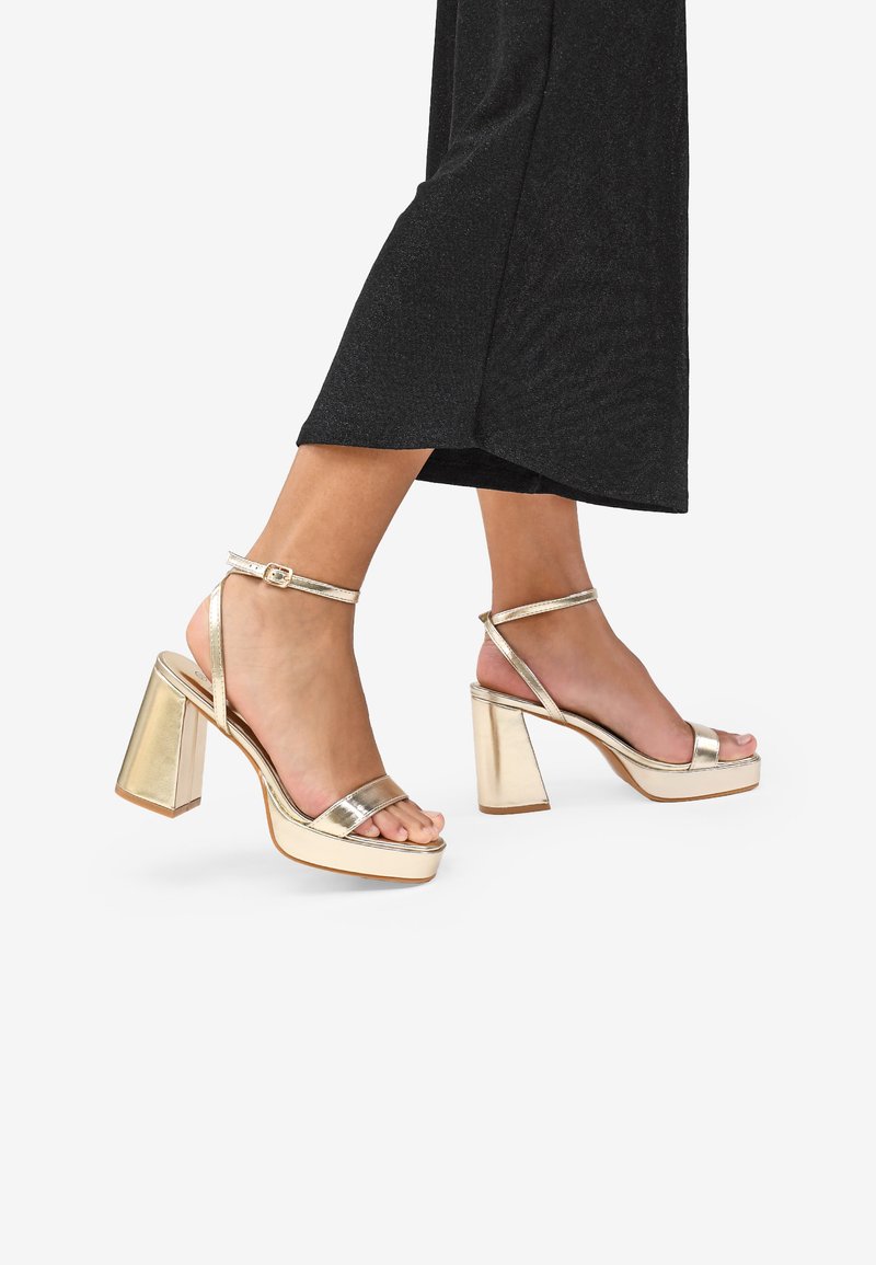 Sandalias de tacón alto metálicas doradas con tacones de bloque, diseño de punta abierta, correa en el tobillo y un acabado suave. Pie parcialmente visible, llevadas con una prenda negra.