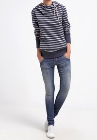 Mikina s námořnickými pruhy a nastavitelnou šňůrkou, přeložená přes jednobarevný námořnický top, spárovaná s modrými skinny džínami a bílými teniskami.