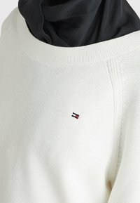 Krämmig stickad sweatshirt med texturerad tyg, ribbade ärmar och en svart huvtröja underneath. Har en liten broderad logotyp i rött, vitt och blått.