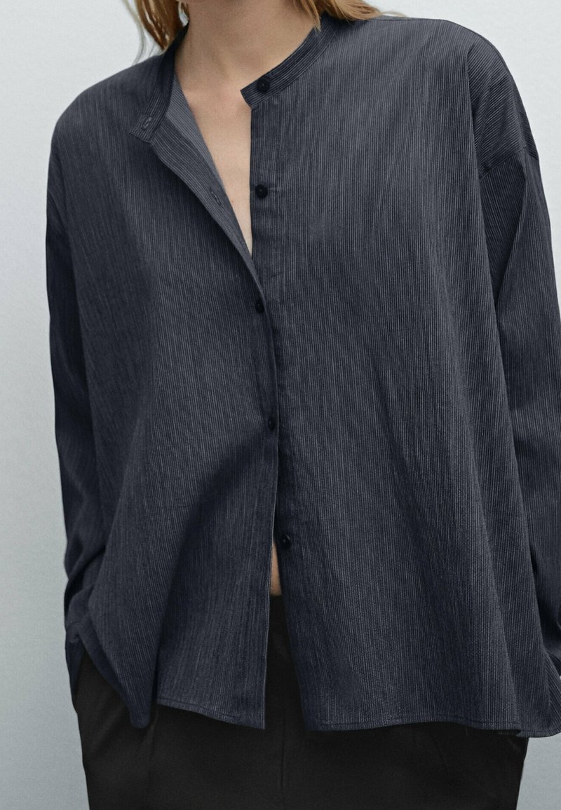 Personne portant une chemise ample gris foncé à rayures verticales avec col mandarin, les mains dans les poches d’un pantalon noir.