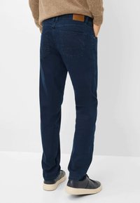 Jean bleu marine au design classique, jambe droite, couture discrète et un patch en cuir beige à la taille. Portés avec des baskets sombres.