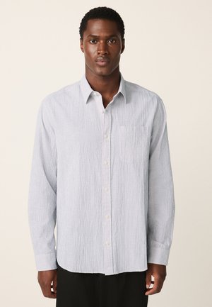 Mees, kes kannab helesinist pika varrukaga nööpidega särki, millel on õrnad vertikaalsed triibud, seisab lihtsa beeži tausta ees.