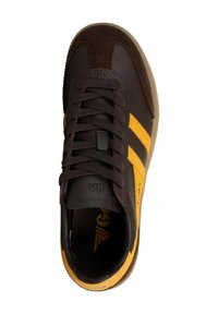 Zapatilla deportiva marrón con franjas de ante amarillo, presenta un upper texturizado, cordones planos y una suela de caucho. Logo interior visible.