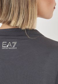 T-shirt grigio scuro con texture a coste, caratterizzato da un logo metallico di "EA7 EMPORIO ARMANI" sul colletto posteriore. Design semplice e moderno.