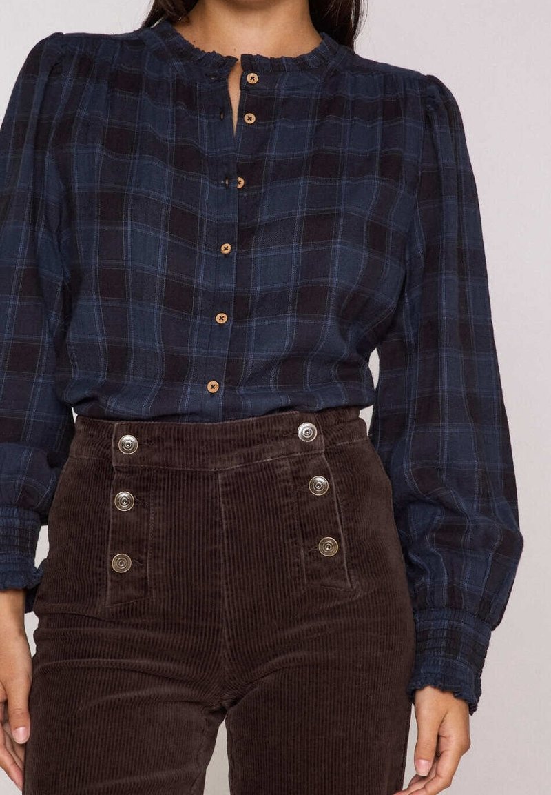 Femme portant un chemisier à carreaux bleu foncé boutonné avec manches longues et un pantalon en velours côtelé marron taille haute avec boutons décoratifs.
