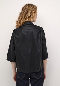 Melna mākslīgā āda, pārāk liela jaka ar trim ceturtdaļām piedurknēm un izgrieztu apkakli. Gluda tekstūra ar taisnu apakšmalu un minimālu apdari.