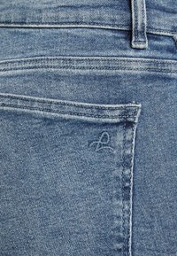 Světle modré džíny z denimu s zadní kapsou, viditelnými švy a malým vyšitým logem na kapse. Měkká textura a klasický střih.