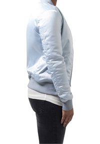 Jasnoniebieska kurtka typu bomber o błyszczącym wykończeniu, z prążkowanymi mankietami i dołem oraz bocznymi kieszeniami. Noszona na białej koszuli i czarnych dżinsach.