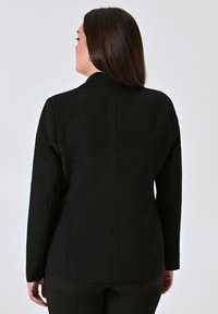 Blazer nero su misura con un design strutturato, caratterizzato da un colletto a lancia e una texture liscia. La vista posteriore mostra una silhouette aderente e linee pulite.