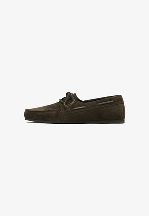 Donkerbruine suède loafer met mocassin-stiksels, veterstrik bovenop en lage zwarte zool, zijaanzicht op witte achtergrond.