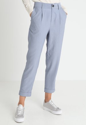 Hellblaue, maßgeschneiderte Hose mit hochgekrempelten Säumen über silbernen Schnürschuhen an einer Person, die mit den Händen in den Taschen steht.