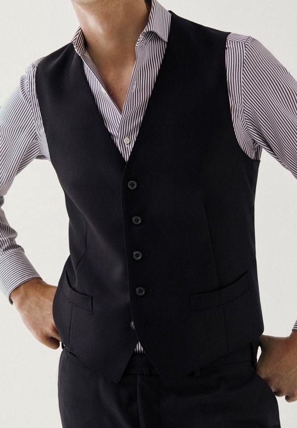 Mand iført en sort knapvest over en hvid og lilla stribet langærmet skjorte, stående med hænderne på hofterne.